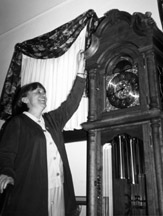 Photo of Peggy Elliott standing next to a grandfather clock reaching up to feel the woodwork.