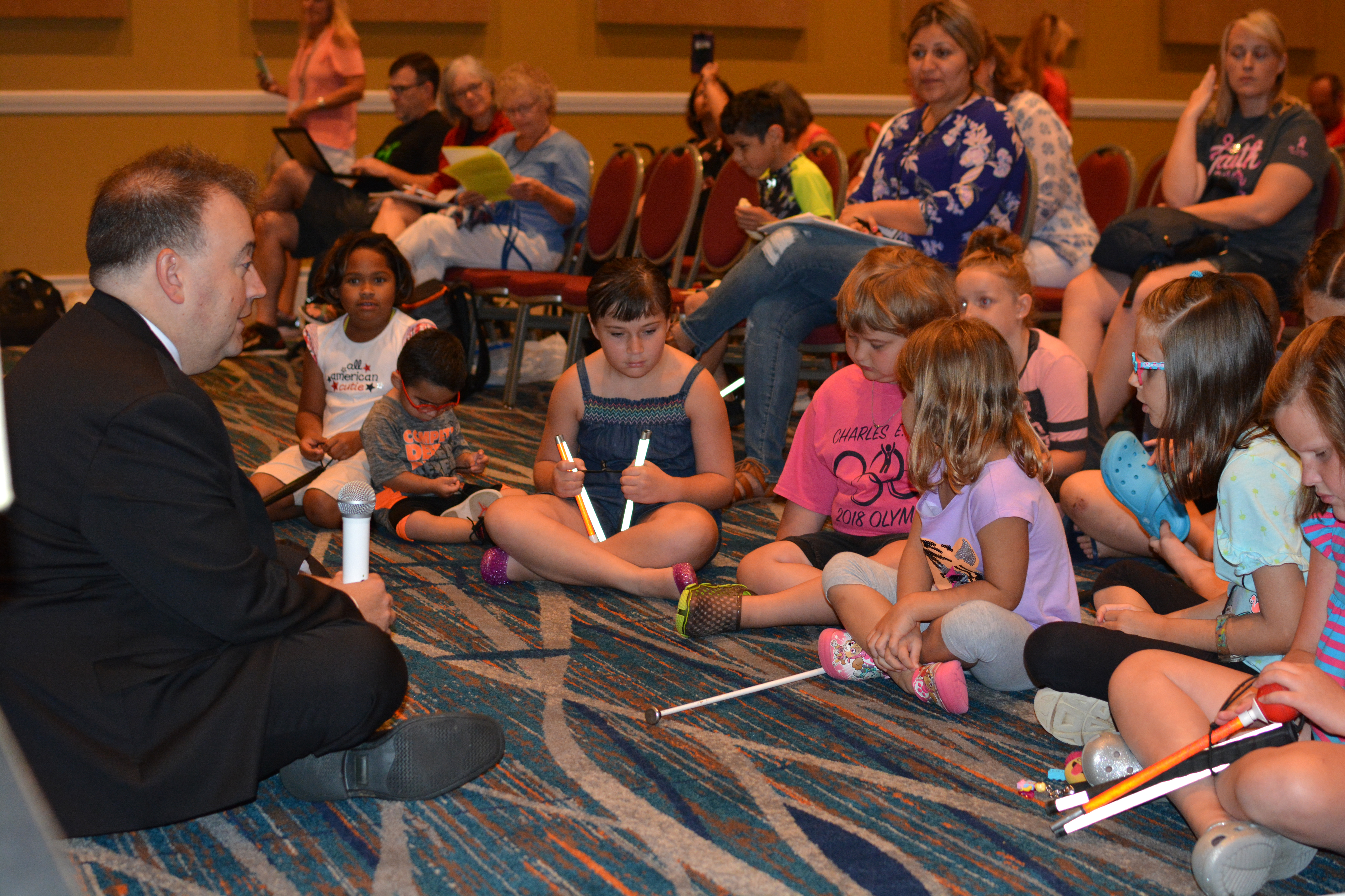 At the NOPBC Conference, NFB President Mark Riccobono sits on the floor surrounded by children.